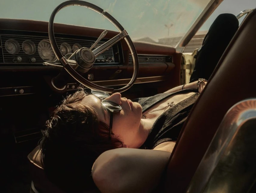 Adult laying back in the driver's seat of a classic car with a steering wheel, dashboard, and outdoor background during daylight.