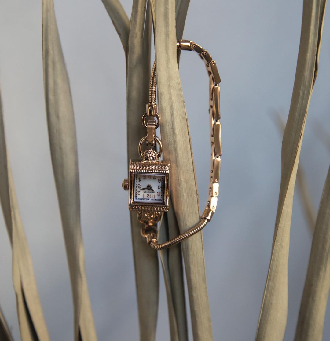 Close-up of a vintage square-faced gold wristwatch with a decorative band, nestled among tan, dried plant fronds, on pale blue background.