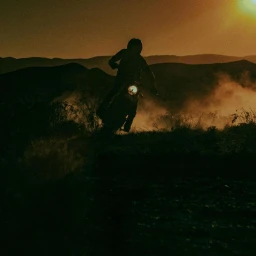 Silhouette of a person riding a motorcycle kicking up dust across a hilly landscape during sunset.