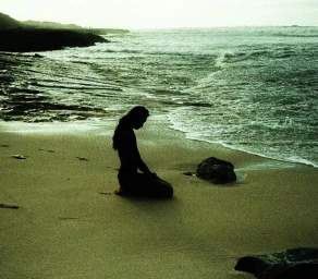 Silhouette of a person kneeling on a sandy beach by breaking ocean waves under an overcast sky captured by Sebastien Zanella.