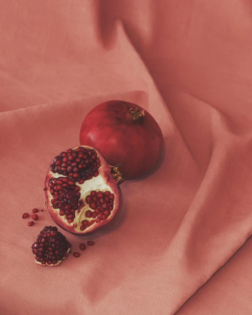 Still life photograph of a whole pomegranate, a halved pomegranate and seeds on a peach-colored fabric with soft lighting.