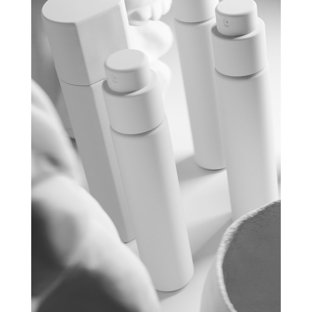 Monochrome shot of several white cosmetic containers, including spray bottles and a bowl, set on a white surface with soft, diffused lighting.