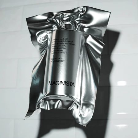 Product shot of a silver foil-wrapped bottle of Maginista hair care product against a white tiled wall with soft, diffused lighting.