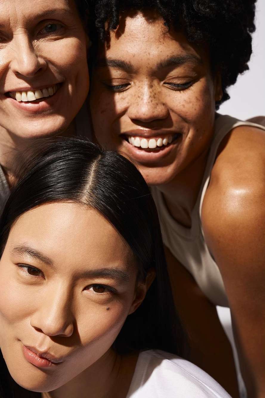 Close-up of three smiling women of diverse ethnicities in a studio setting, showcasing natural beauty and skincare.
