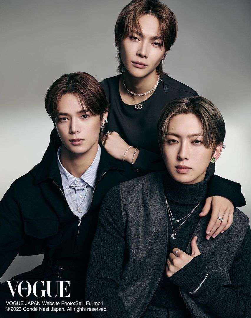 Portrait of three young Asian men in formal dark clothing, stylish necklaces, and earrings, posed in a photo studio for Vogue Japan.
