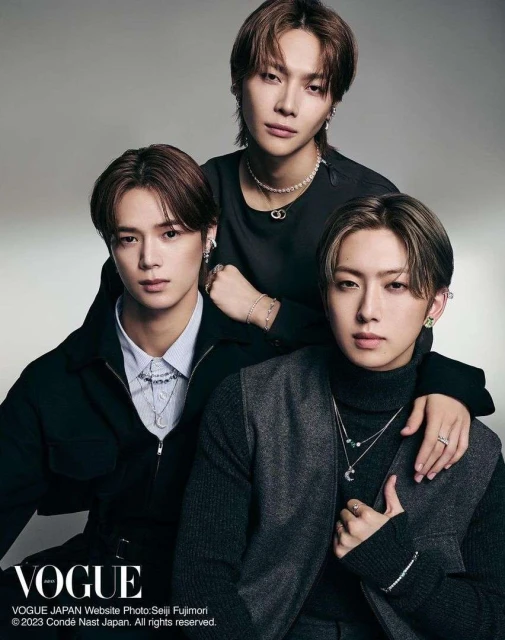 Portrait of three young Asian men in formal dark clothing, stylish necklaces, and earrings, posed in a photo studio for Vogue Japan.