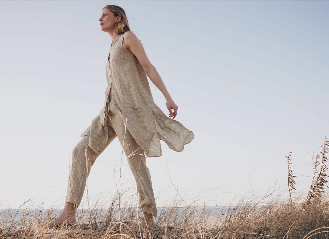 Adult female model walking on a beach in linen clothing with a blue sky background and neutral color palette, outdoors in daylight.