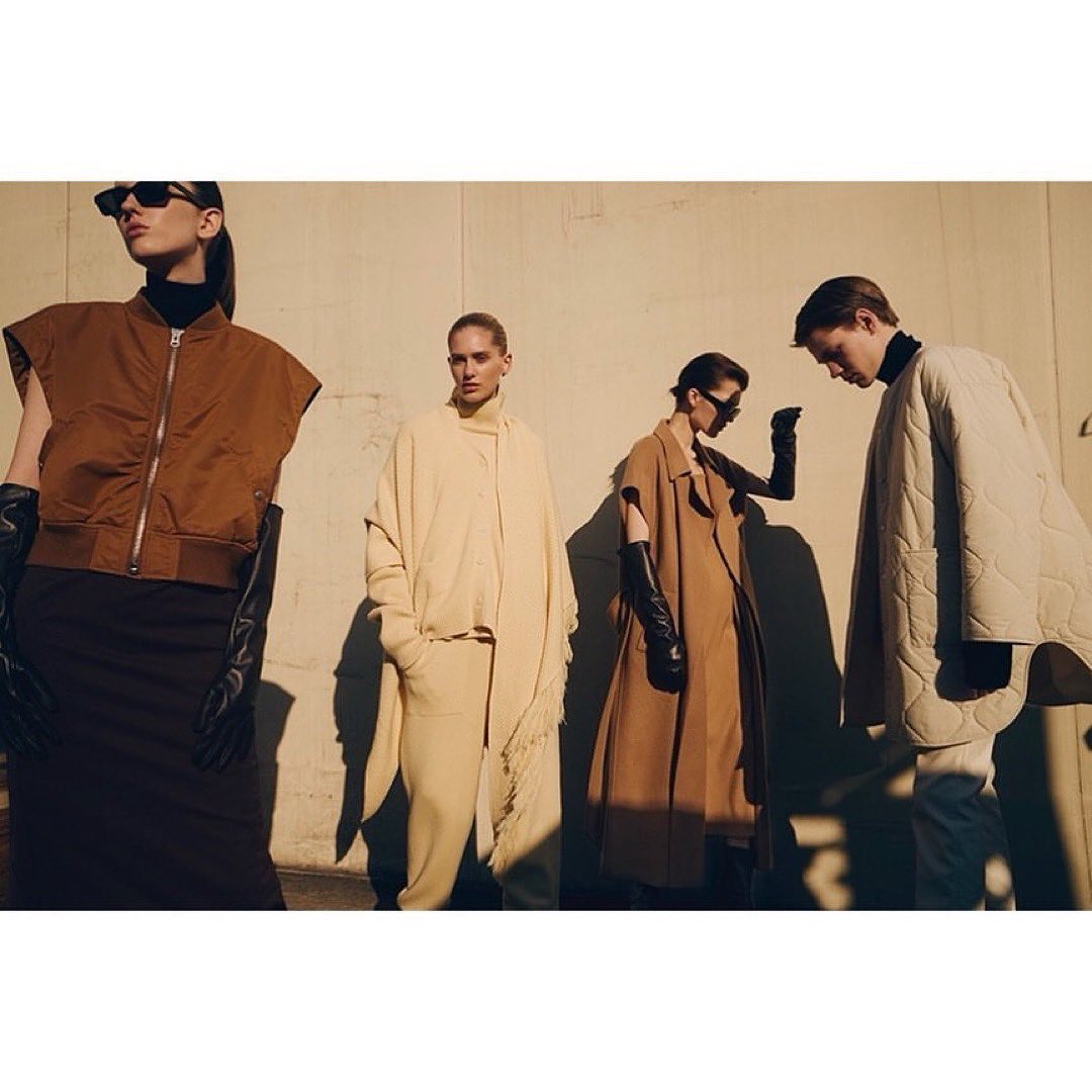 Four fashion models in neutral-tone outfits posing in front of a cream-colored wall, wearing sunglasses and long gloves in a studio setting.