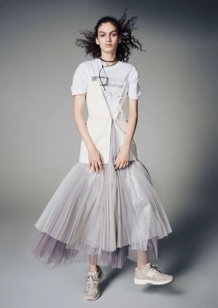 Model with dark curly hair wearing a white t-shirt, asymmetric layered skirt, and sneakers against a plain grey background