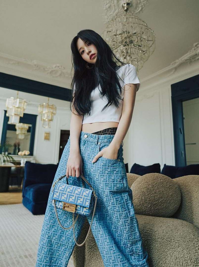 A brunette woman in patterned jeans holds a small blue handbag in an indoor setting with a chandelier and couch.