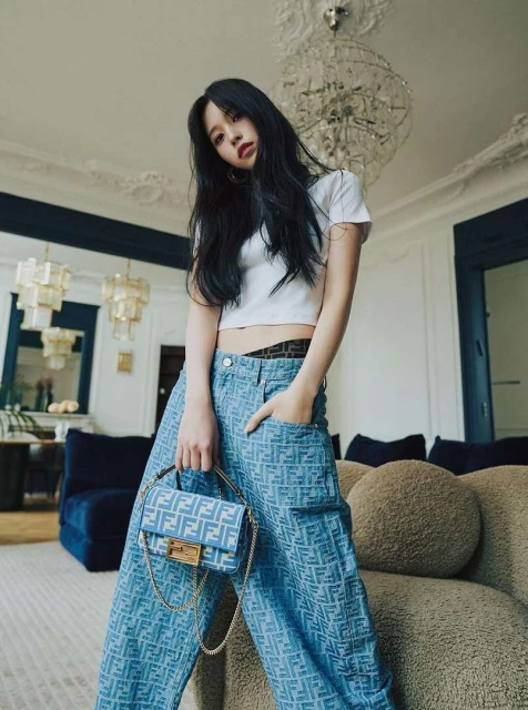 A brunette woman in patterned jeans holds a small blue handbag in an indoor setting with a chandelier and couch.