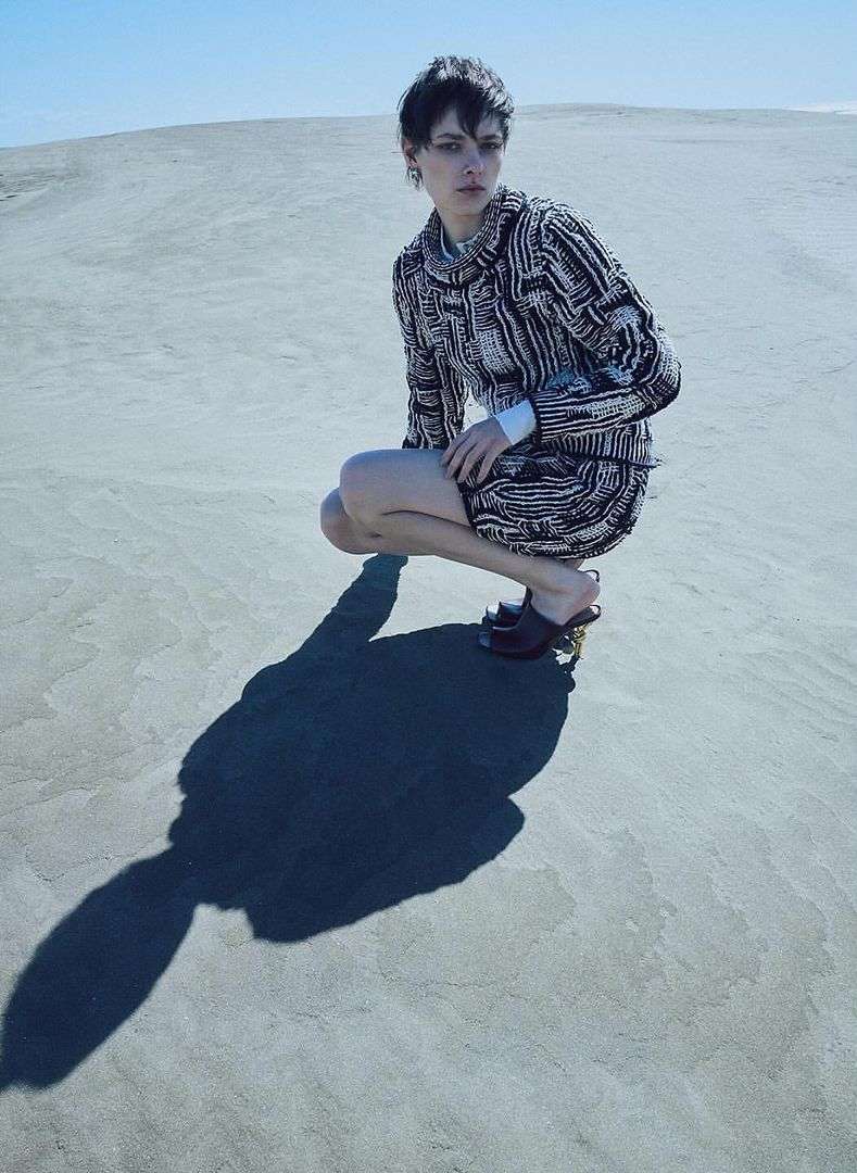 Model with short dark hair crouching in sand dunes wearing a patterned textured sweater and dark heeled shoes by Seiji Fujimori