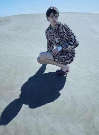 Model with short dark hair crouching in sand dunes wearing a patterned textured sweater and dark heeled shoes by Seiji Fujimori