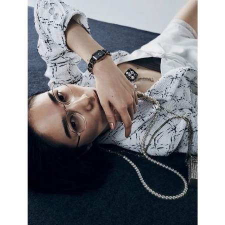 Woman in tweed jacket and round glasses reclining on textured floor with pearl necklace and watch