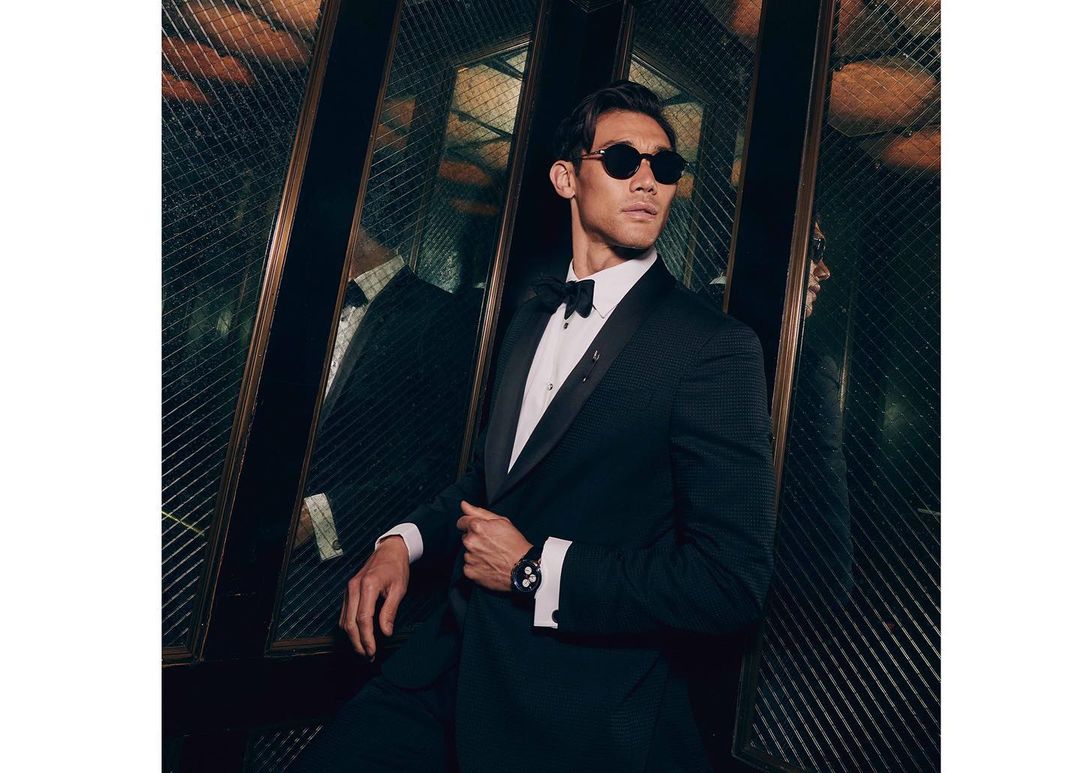 Portrait of stylish adult male in a dark suit and bow tie leaning against a patterned wall with sunglasses, shot indoors with dark lighting.
