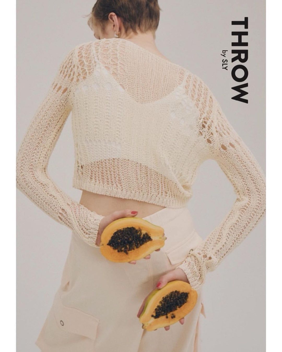 Back view of an adult woman wearing a beige open-knit sweater and holding two halved papayas in a studio setting on white backdrop.