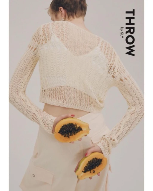 Back view of an adult woman wearing a beige open-knit sweater and holding two halved papayas in a studio setting on white backdrop.