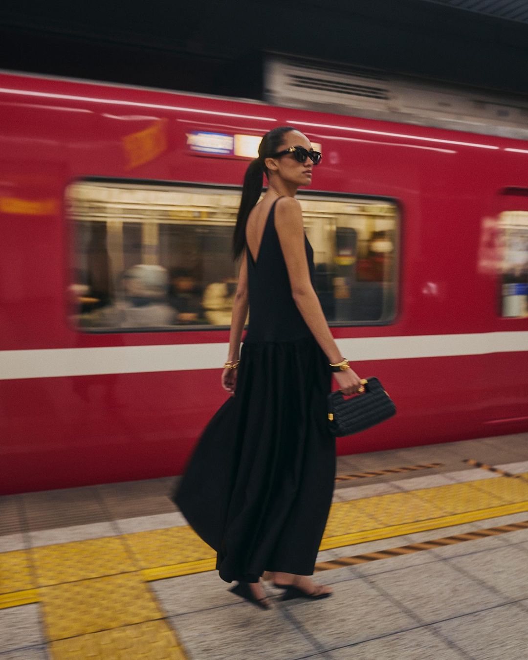 Woman in a black dress walks along a subway platform with a red train blurred in motion behind her, accessorized with jewelry and a handbag.