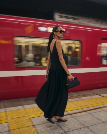 Woman in a black dress walks along a subway platform with a red train blurred in motion behind her, accessorized with jewelry and a handbag.