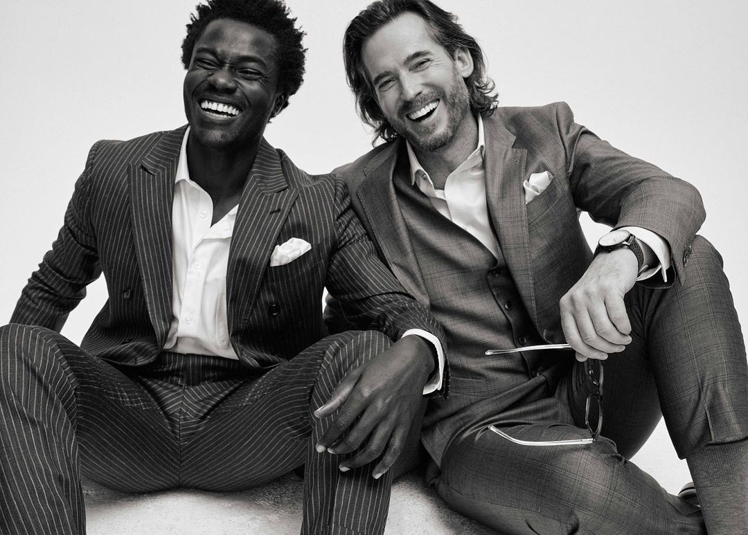 Two smiling men in suits sit together. The African-American man is in a pinstripe suit, the Caucasian man in a gray suit.