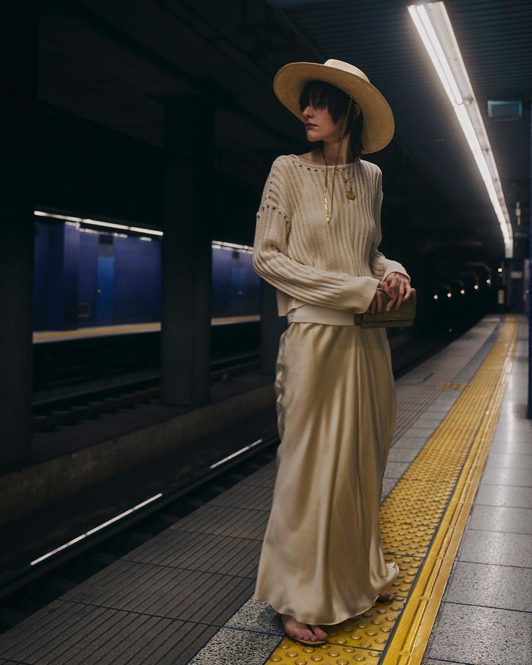 Model wearing a wide-brim straw hat, ribbed cream sweater, and silk maxi skirt standing on a subway station platform.