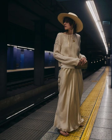 Model wearing a wide-brim straw hat, ribbed cream sweater, and silk maxi skirt standing on a subway station platform.