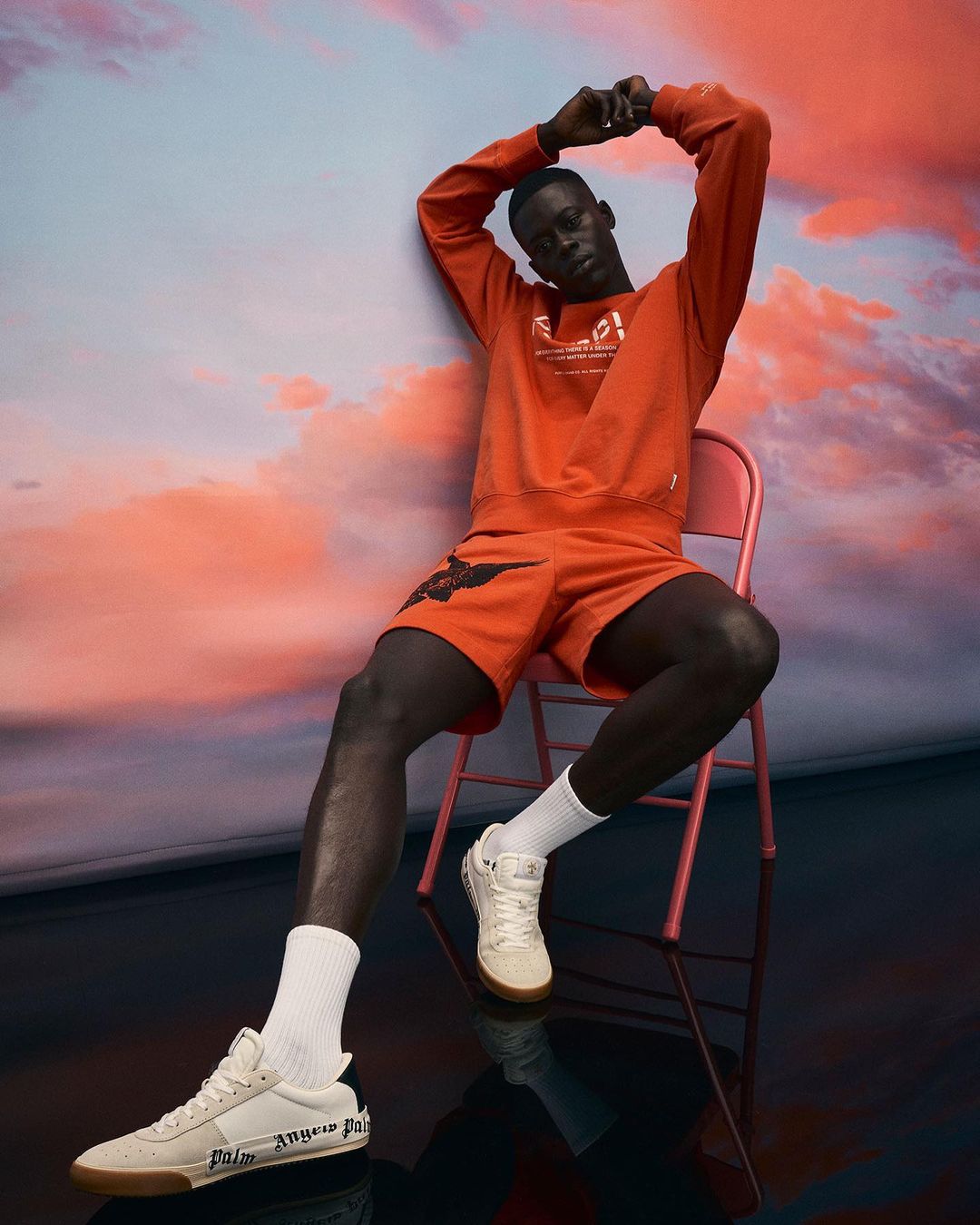 An adult male model poses while sitting on a pink chair in orange matching sweatshirt and shorts with a sunset backdrop, wearing sneakers.