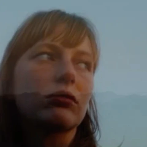 Portrait of fair-skinned adult female with parted blonde hair and dark lipstick, looking up against a soft blue sky and mountain.