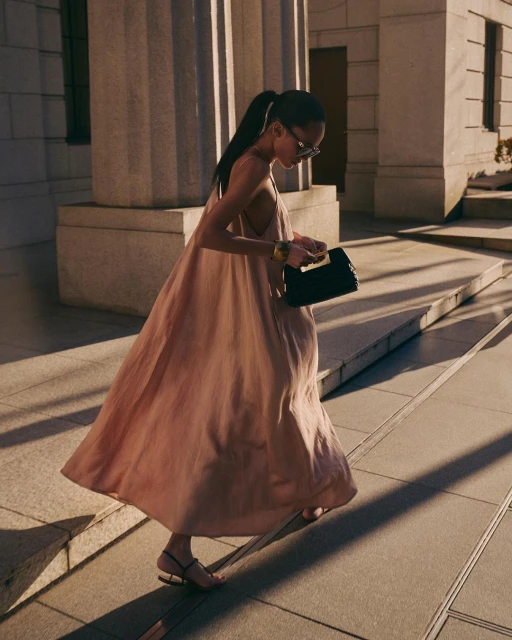 Woman in flowing dress ascends stone steps carrying black clutch, accessorized with bracelet and sunglasses in urban setting.