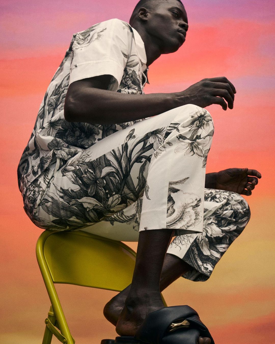 An adult Black male poses on a yellow metal chair. He is wearing a matching black and white botanical print outfit with black slides.
