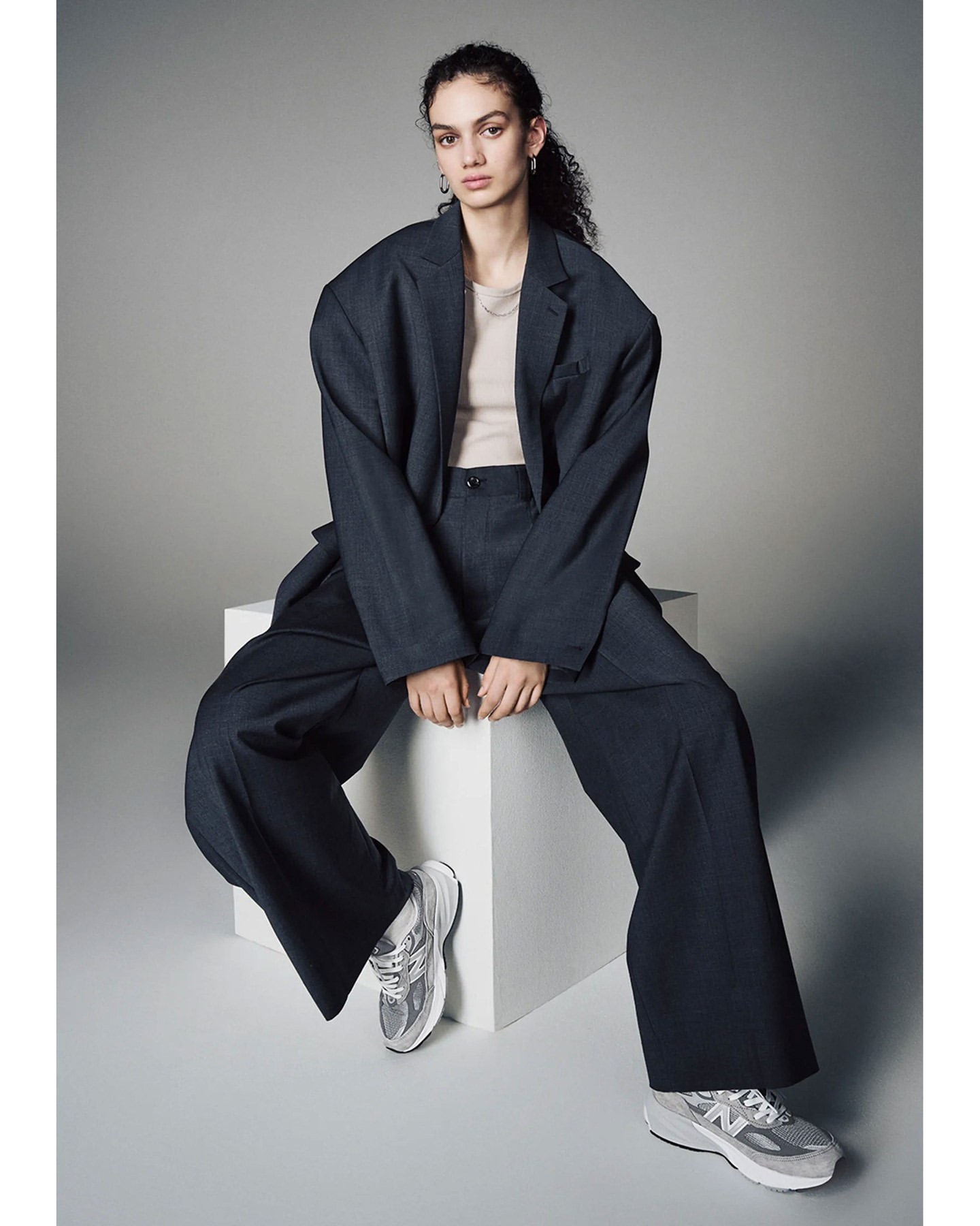 A woman sits in a dark suit with wide-legged pants and silver sneakers, posed against a light gray background in a studio setting.