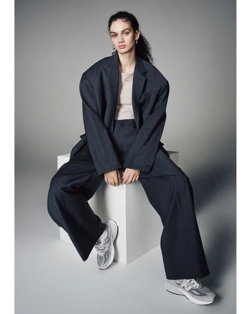 A woman sits in a dark suit with wide-legged pants and silver sneakers, posed against a light gray background in a studio setting.