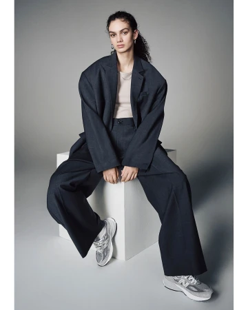 A woman sits in a dark suit with wide-legged pants and silver sneakers, posed against a light gray background in a studio setting.