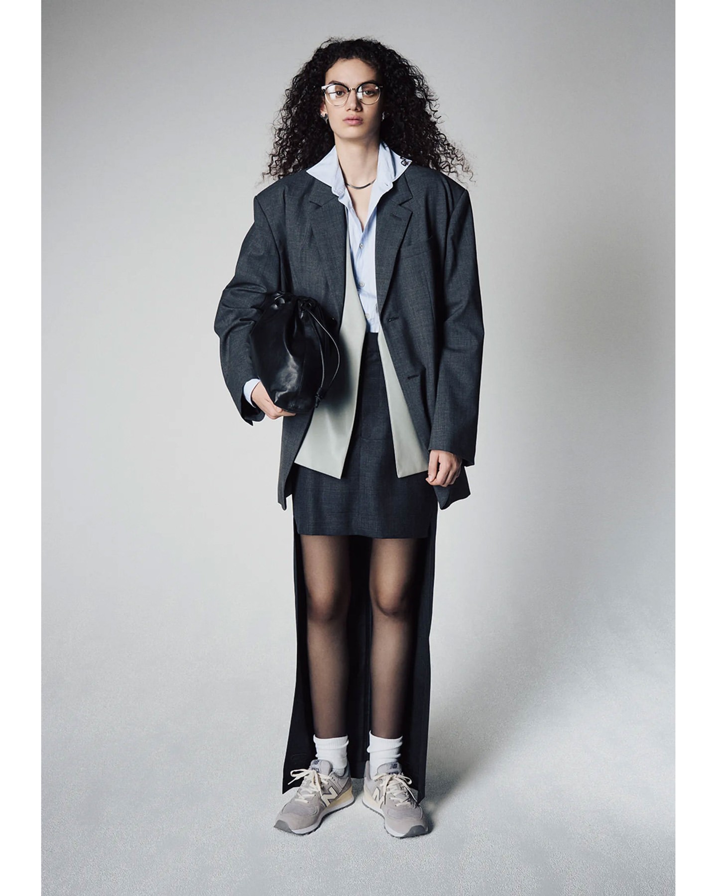 Woman in oversized gray blazer and skirt, wearing sneakers, holds a black purse in a studio setting with a light gray background.