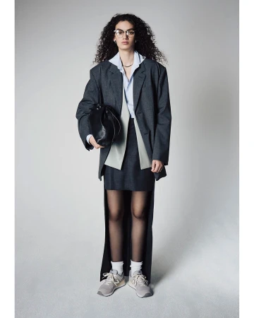 Woman in oversized gray blazer and skirt, wearing sneakers, holds a black purse in a studio setting with a light gray background.
