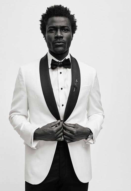 Portrait of a Black male model in a white tuxedo jacket with a black bow tie looking at the camera in a photo studio with neutral lighting.
