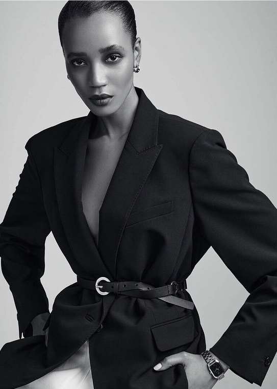A stylish woman poses in a black blazer cinched with a belt, complemented by a wristwatch, against a plain background.