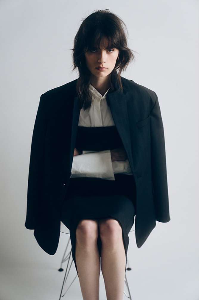 Model seated on a chair wearing a dark blazer over a white shirt against a plain white background