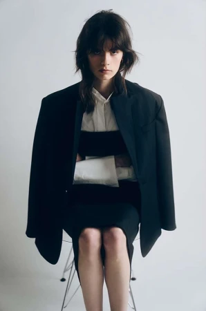 Model seated on a chair wearing a dark blazer over a white shirt against a plain white background