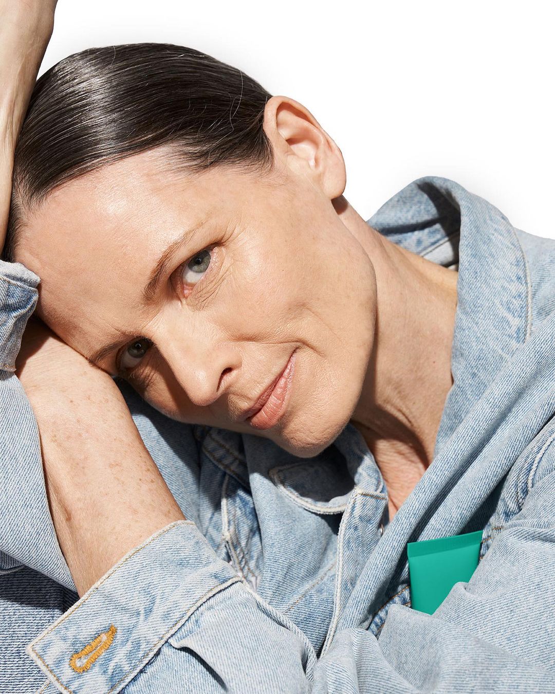 Headshot of a middle-aged woman with slicked-back dark hair, wearing a denim jacket, and leaning her head on her arms against a white backdrop.