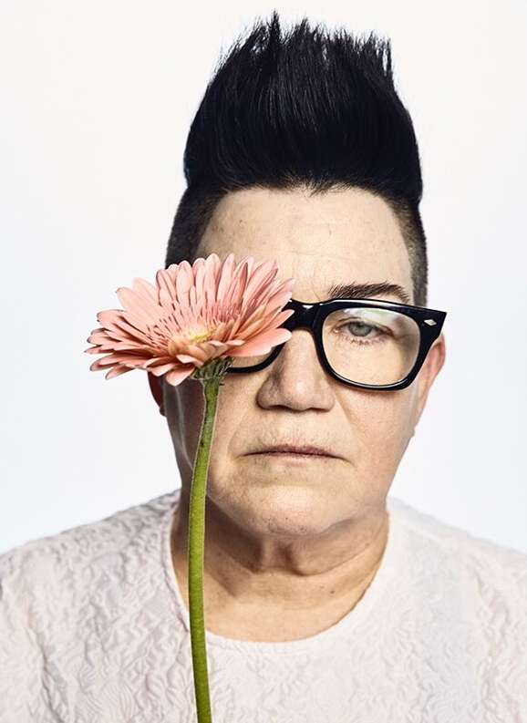 Headshot of a person with a high pompadour hairstyle and glasses, holding a pink gerbera daisy in front of their face on a white background.