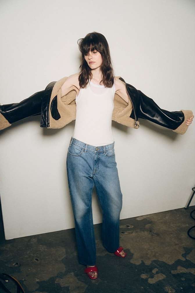 Brunette adult woman wearing a white top, jeans, and a brown and black jacket in front of a white studio wall. 