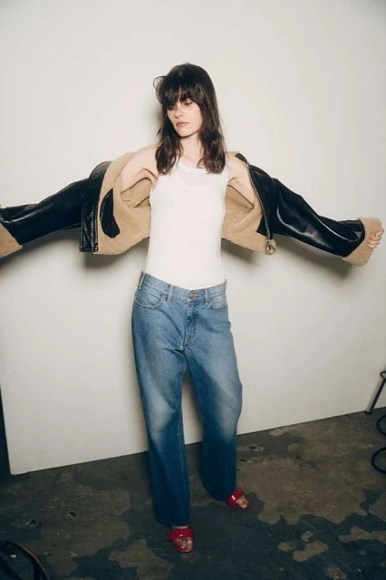 Brunette adult woman wearing a white top, jeans, and a brown and black jacket in front of a white studio wall. 