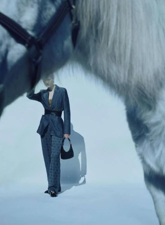 Model in a patterned blue suit holding a handbag framed between the legs of a white horse in a studio