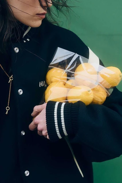 Woman in a black varsity jacket holds a clear bag full of lemons against a green backdrop, showcasing a blend of fashion and food.
