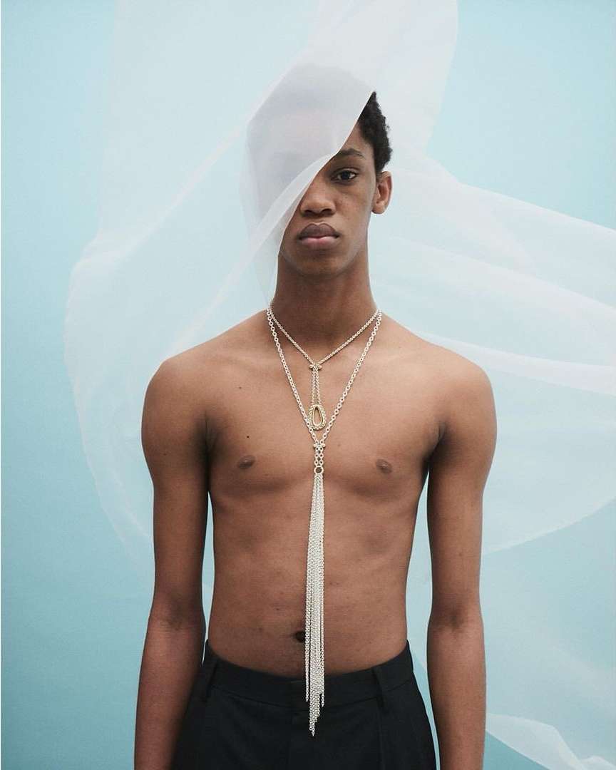 Studio shot of a topless male model with a sheer white fabric and a long beaded necklace against a pale blue backdrop. 