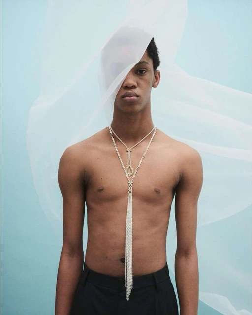 Studio shot of a topless male model with a sheer white fabric and a long beaded necklace against a pale blue backdrop. 