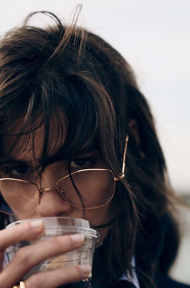 Close-up of a young adult woman with brown hair and eyeglasses drinking from a plastic cup outdoors on an overcast day.