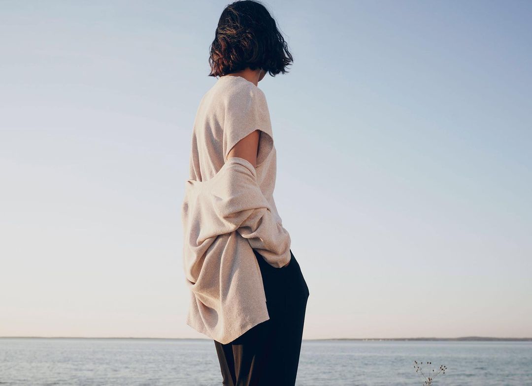 Woman standing outdoors near the ocean wearing a beige sweater and dark pants enjoying the serene view on a bright day.