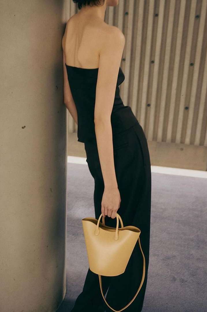 A woman in a black strapless top and trousers poses against a grey wall, holding a tan leather handbag with handles.